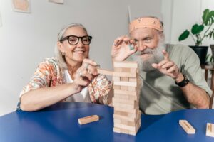 Para seniorów, kobieta i mężczyzna, układają wieżę z drewnianych klocków przy stole, ciesząc się wspólną zabawą.